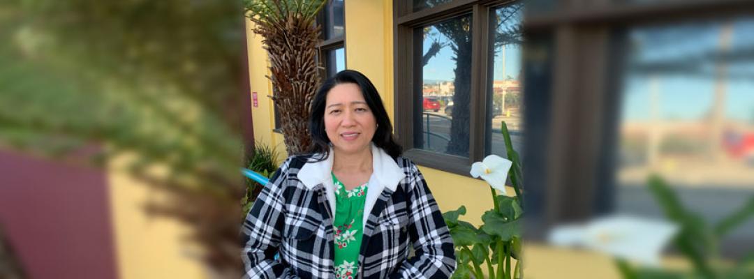 Carmel Gacho in plaid jacket in front of a tree and yellow building