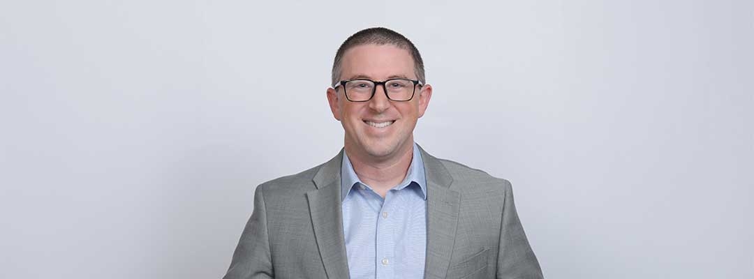 Photo of Elliott Asarch wearing a suit standing in front of a gray background