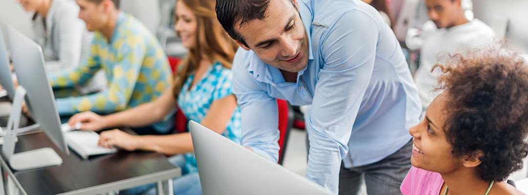 Photo of adult teacher leaning over to speak with a student who is sitting at a laptop