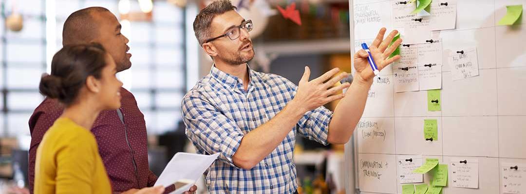 Man pointing at whiteboard with sticky notes while 2 colleagues look on