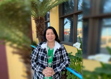 Carmel Gacho in plaid jacket in front of a tree and yellow building