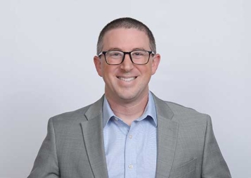 Photo of Elliott Asarch wearing a suit standing in front of a gray background