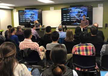 Audience seated and watching a presentation from moderator Sheng Huang in front of a plasma display