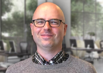 Christopher Hubbard posing while wearing a cardigan over a plad button-down shirt in front of large windows and computer stations  