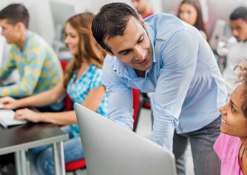 Photo of adult teacher leaning over to speak with a student who is sitting at a laptop