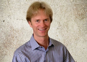 Mark Schrier posing in front of a concrete wall wearing a striped button down shirt