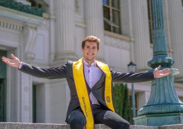 Markus sits atop a ledge in front of Doe Library wearing a suit and the Berkeley graduation stole, smiling with his arms outstretched 